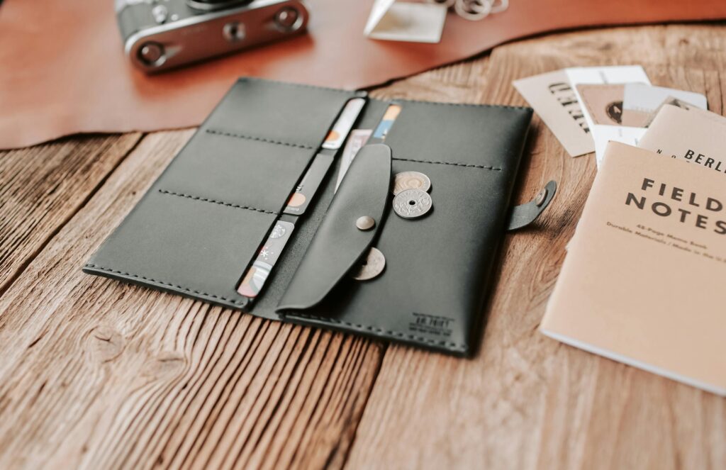 Stylish leather wallet open on a rustic wooden surface with coins, camera, and field notes nearby.